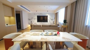 A modern hotel room with a marble-topped dining table, four chairs, a sofa, a TV mounted on the wall, and a small side table with a plant.