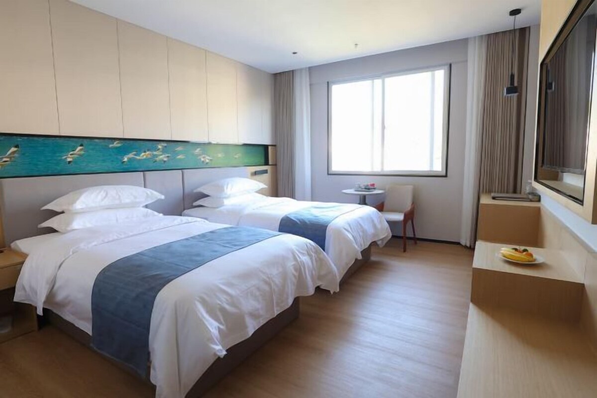 A hotel room with two beds, a small table, a chair, a window with curtains, and a fruit bowl on a side table.