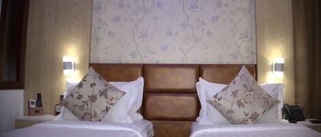 A hotel room with two beds, a wooden headboard, floral wallpaper, bedside lamps, and a nightstand with a telephone and a clock.