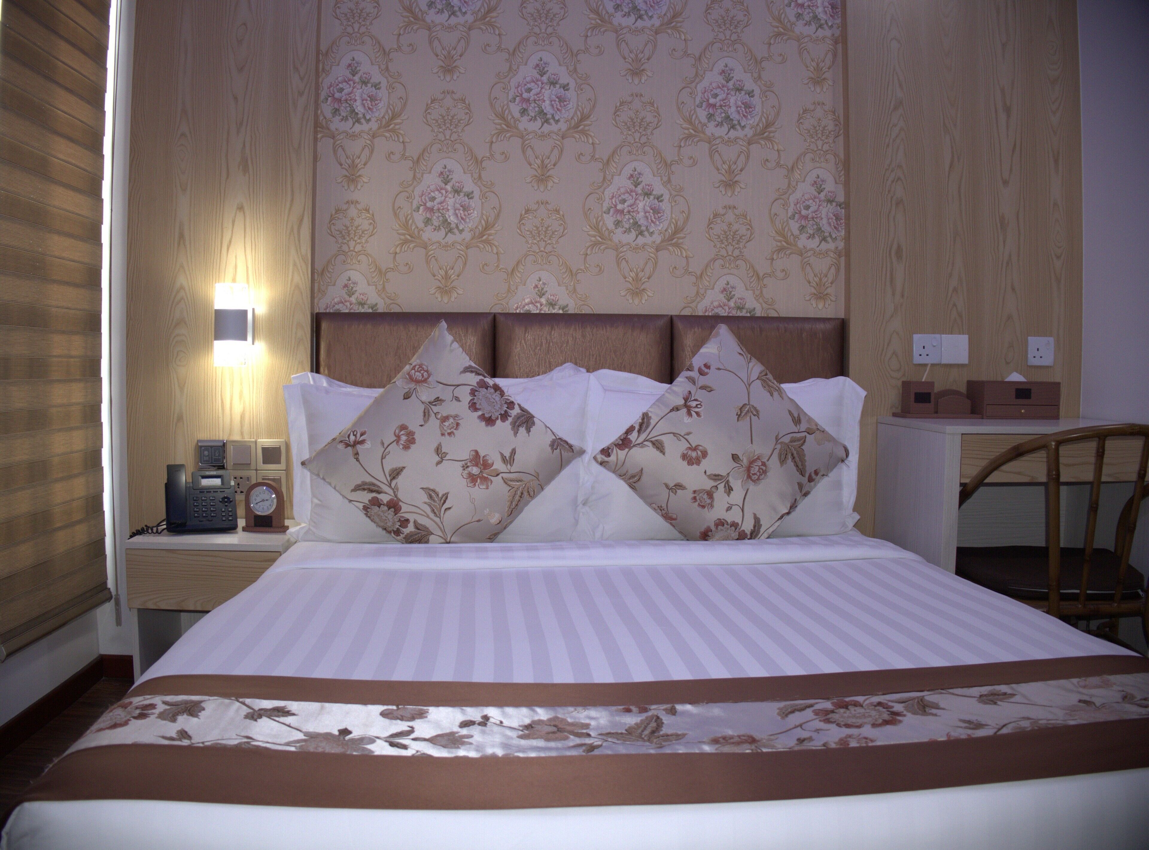 A neatly made bed with floral pillows, a bedside table with a phone and clock, and a desk with a chair in a hotel room.