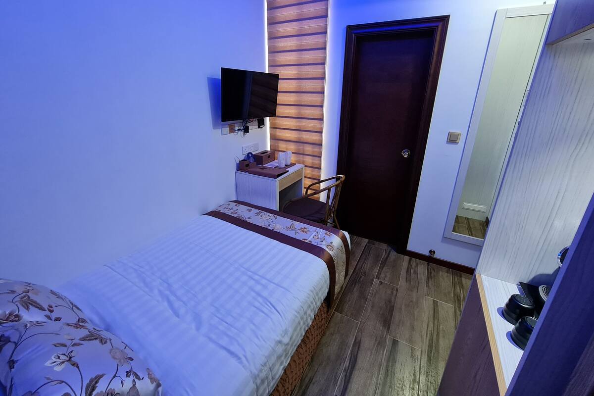 A compact bedroom with a bed, a small bedside table, a chair, a wardrobe, and a television mounted on the wall.