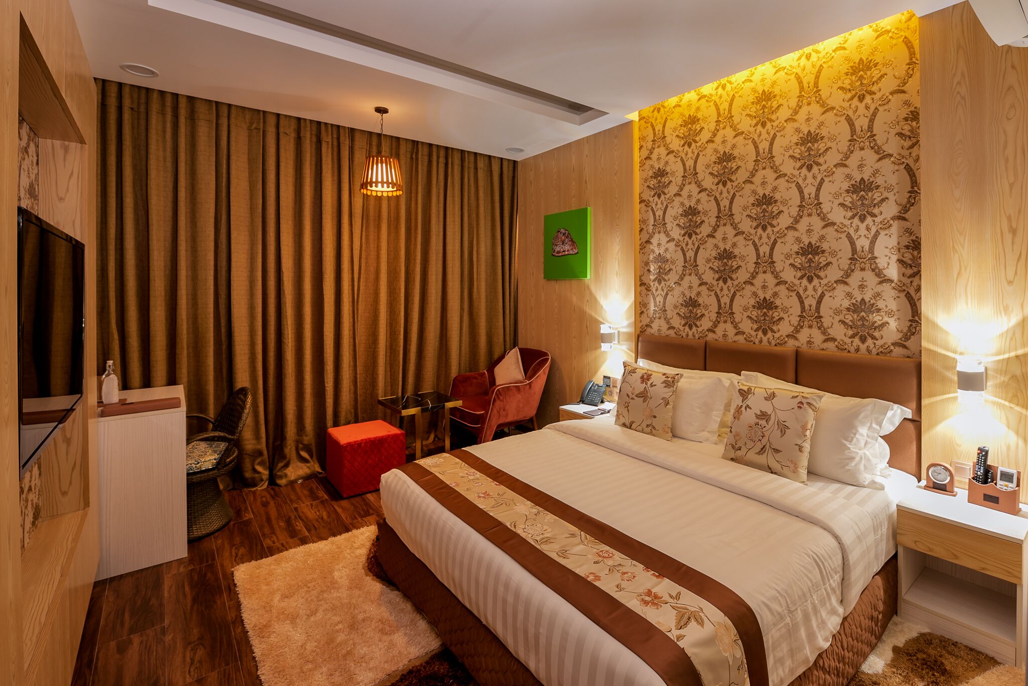 A hotel room with a large bed, a TV, a desk, a chair, and a red ottoman.