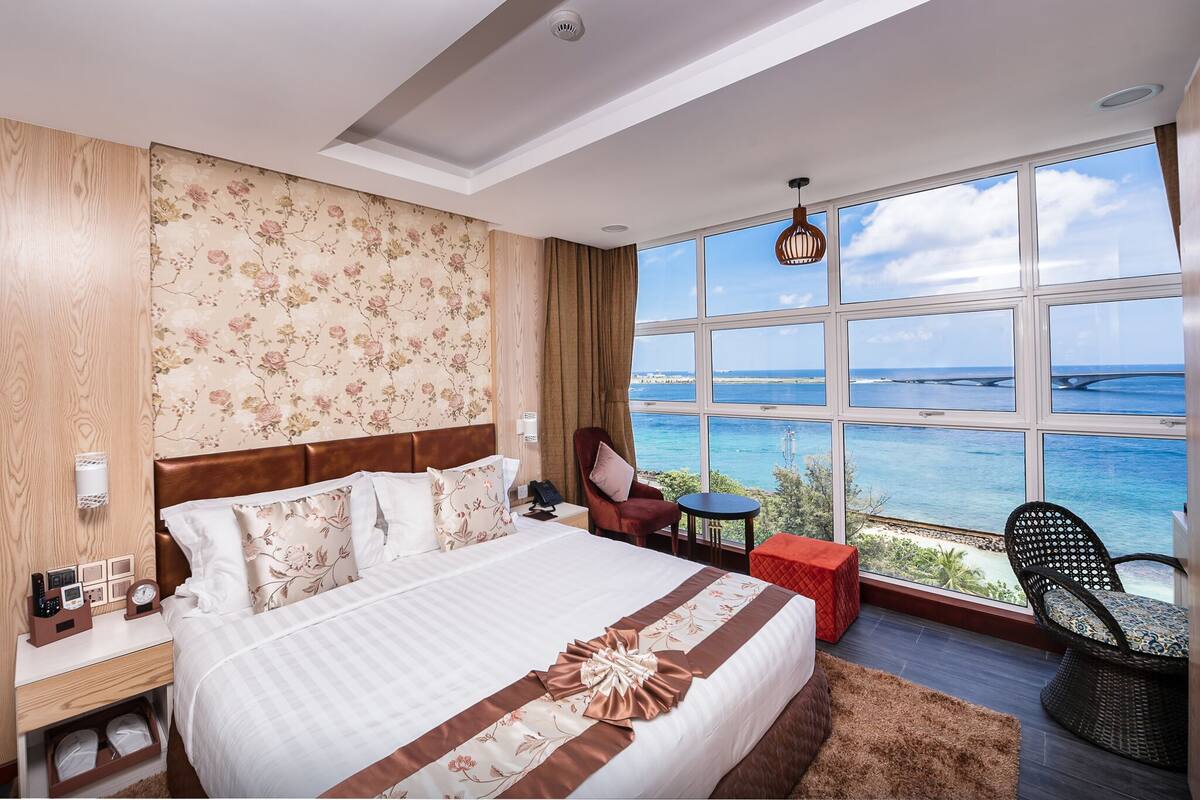 A hotel room with a large bed, a view of the ocean, a wooden headboard, and a small table with a chair.