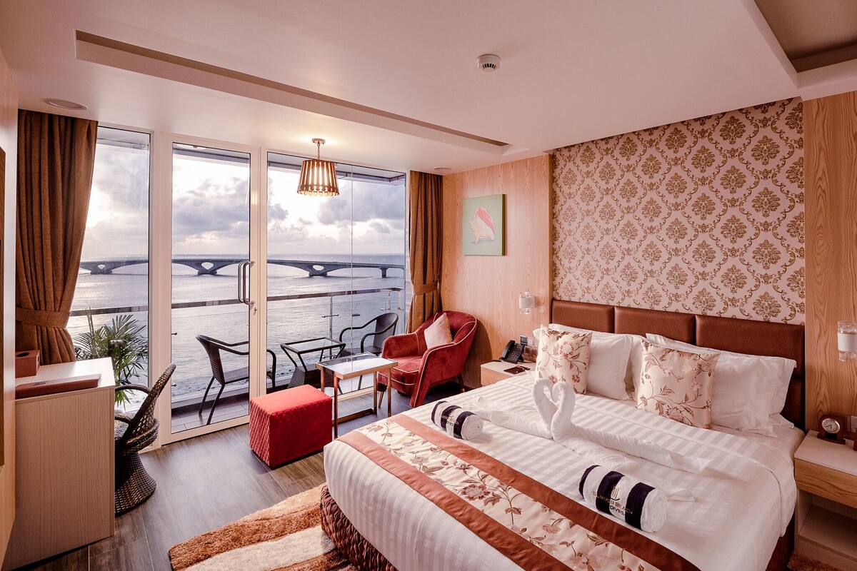 A hotel room with a large bed, a red chair, a small table, and a view of a bridge over water.
