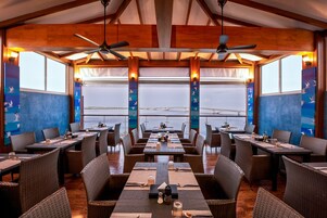 Un restaurante con varias mesas y sillas, amplios ventanales con vistas y vigas de madera en el techo.