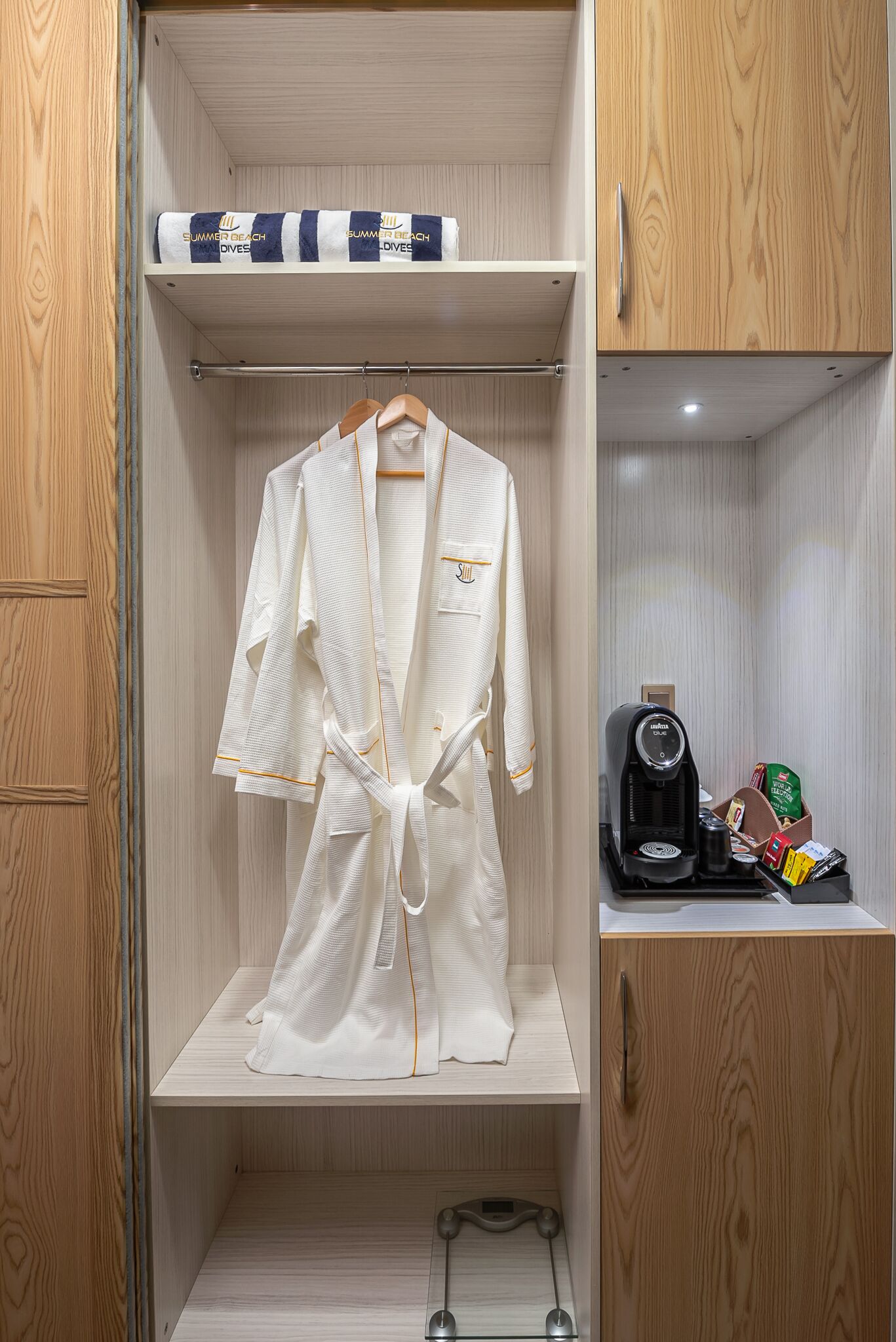 A robe hanging in a wooden closet with a shelf above containing a towel and a box labeled "Summer Beach Maldives."