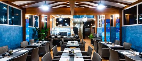 A restaurant with long tables and wicker chairs, wooden ceiling beams, and a blue wall with a mural.
