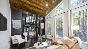 Een moderne woonkamer met een open haard, leren bank en grote ramen met uitzicht op het bos.