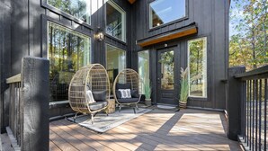 Een modern huis met een houten terras, grote ramen en twee rieten stoelen met kussens.