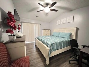 A bedroom with a bed, desk, chair, ceiling fan, and a window with blinds.