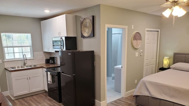 A compact kitchen with white cabinets, a black refrigerator, and a microwave. There is a small dining area with a table and chairs. The room has a bed with a gray bedspread and a nightstand with a lamp. - Sanctuary, Lackland AFB, SeaWorld Fiesta Texas (San Antonio)
