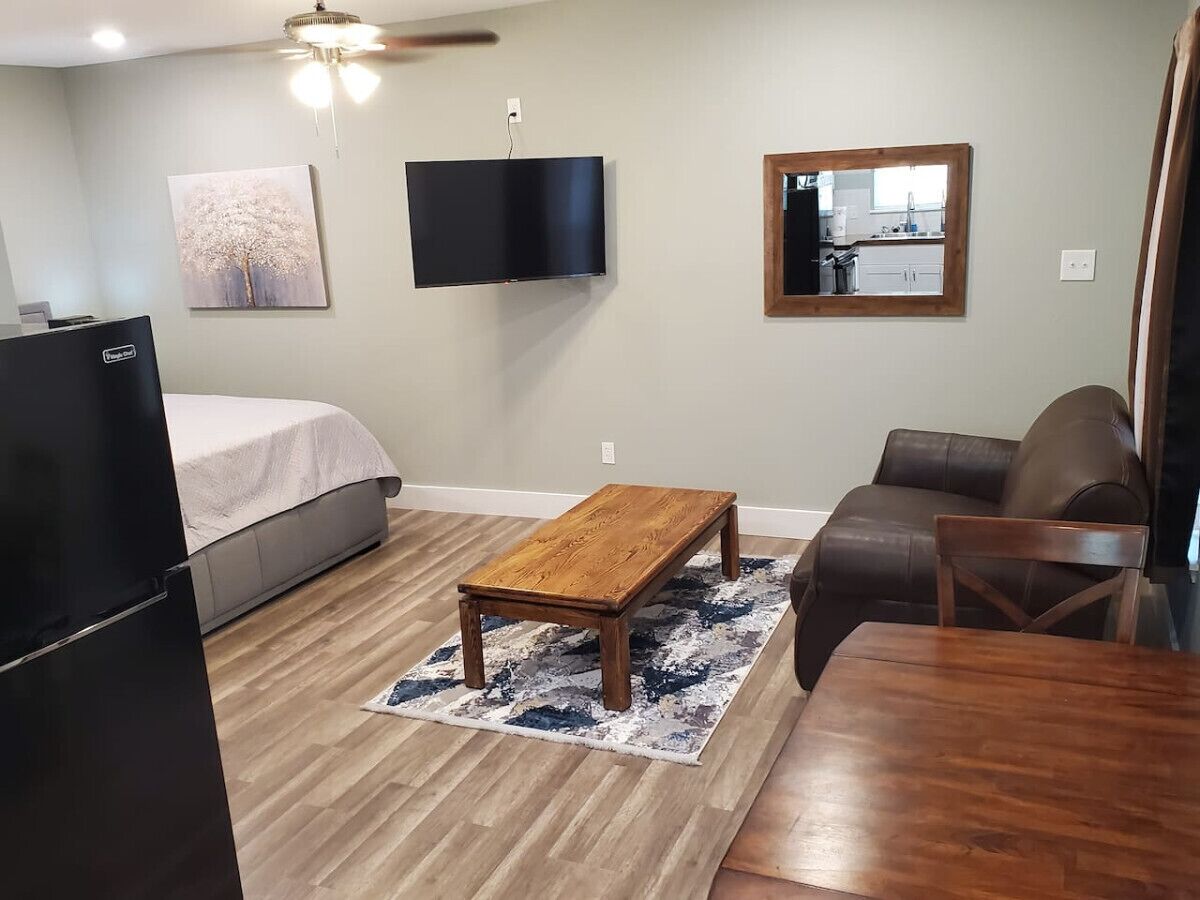 Un dormitorio con cama, una mesita de centro de madera, un sofá de cuero marrón, una mesa de comedor de madera, un televisor montado en la pared y una cocina visible en el reflejo del espejo.