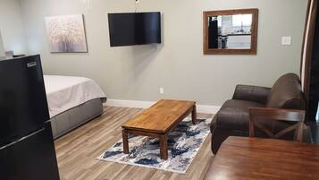 Un dormitorio con cama, una mesita de centro de madera, un sofá de cuero marrón, una mesa de comedor de madera, un televisor montado en la pared y una cocina visible en el reflejo del espejo.
