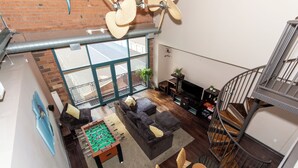 Ein loftsartiger Wohnbereich mit einem Kicker, dunklen Sofas, einem Fernseher und einer Wendeltreppe.
