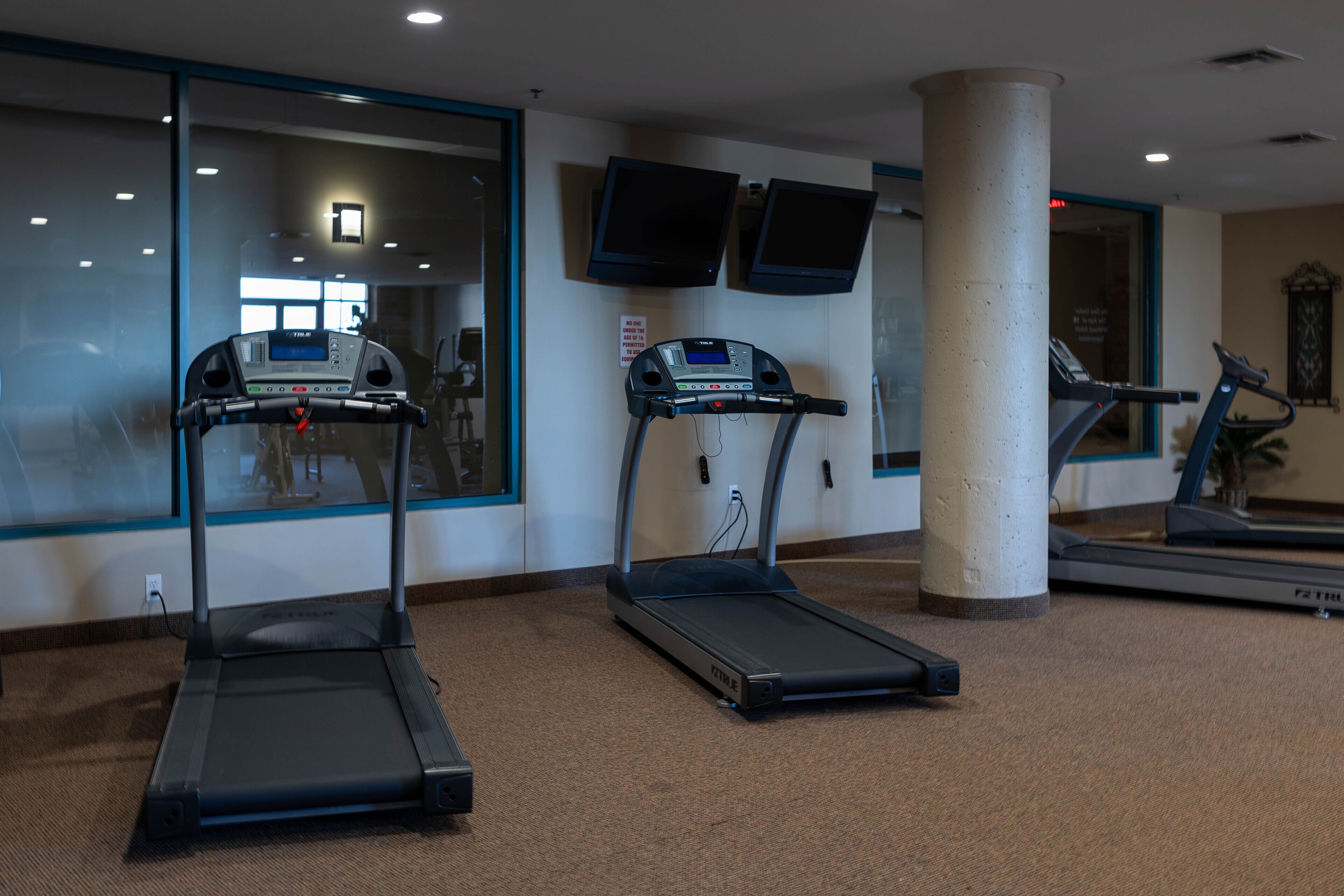 Un gimnasio con cintas de correr, un pilar grande y un televisor instalado en la pared.