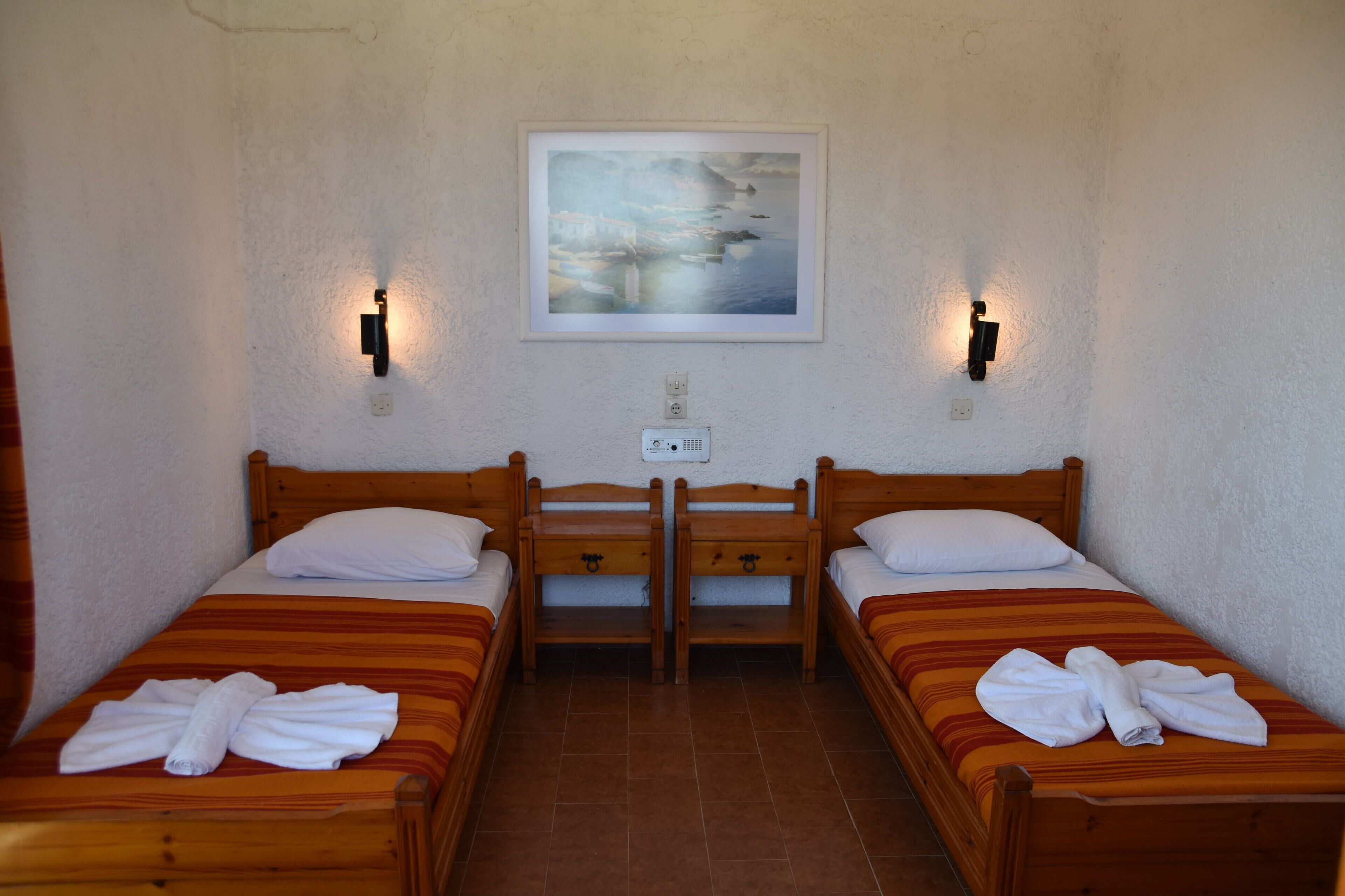 A room with two wooden beds, each with a white pillow and a towel. There is a framed picture on the wall above the beds.