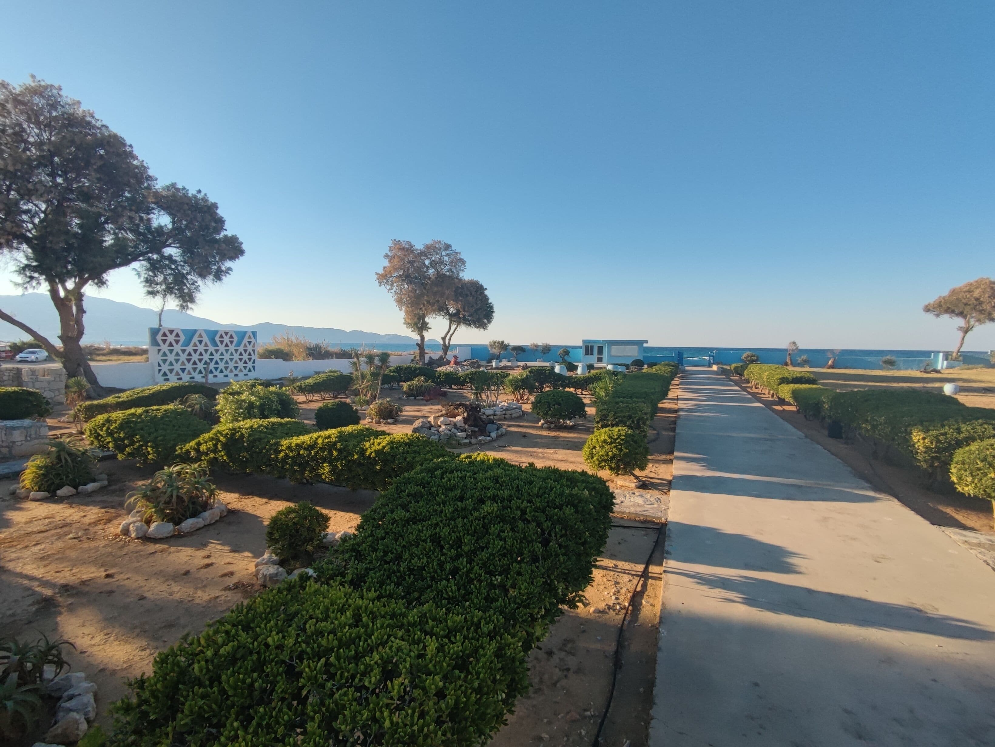 A pathway leading to a building with a sign, surrounded by well-maintained greenery and a clear view of the sea.
