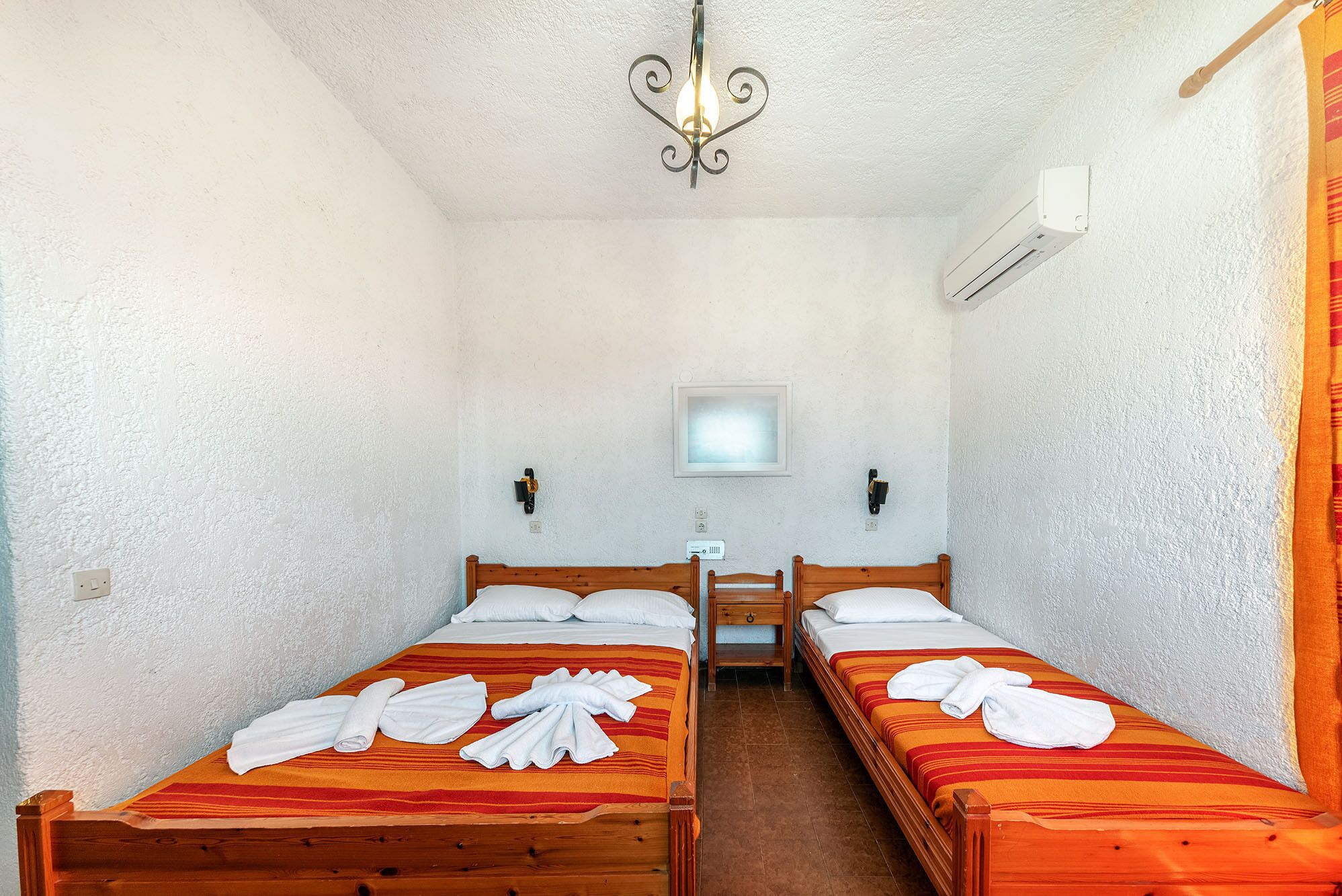A room with two wooden beds, each with a red and orange striped bedspread and white towels. There is a small bedside table with a lamp, a wall-mounted air conditioner, and a ceiling light fixture.