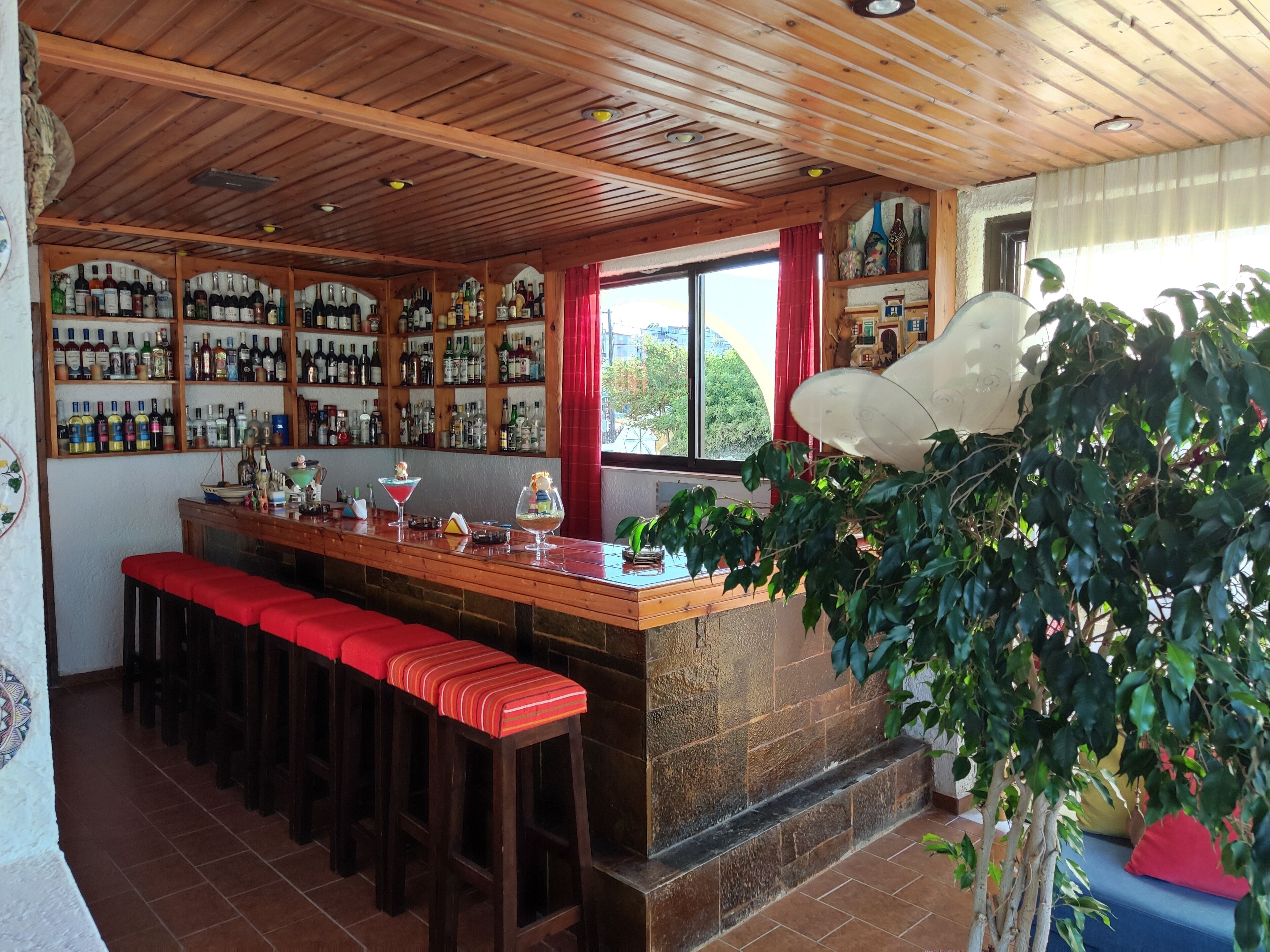 Un espace bar avec des étagères en bois remplies de bouteilles, un long comptoir avec des tabourets et une grande fenêtre ornée de rideaux rouges.