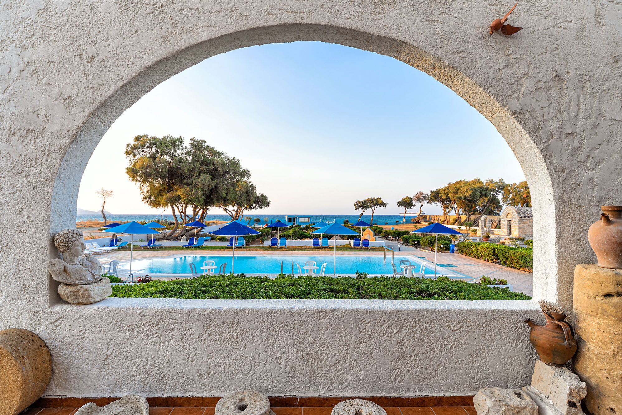 A pool area with blue umbrellas, a clear view of the sea, and a stone archway.