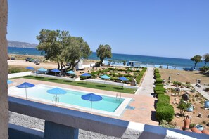 A pool area with blue umbrellas, a clear view of the sea, and a well-maintained garden with neatly trimmed bushes and trees. - Hotel Tsagarakis Beach (Malevizi)