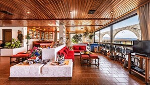 A spacious lounge area with a wooden ceiling, a bar with shelves of bottles, a large window with a view of a bridge, and a seating area with red sofas and a coffee table. - Hotel Tsagarakis Beach (Malevizi)