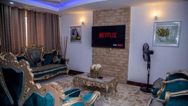 Una sala de estar con una televisión mostrando 'NETFLIX', una pared de ladrillo y un ventilador.