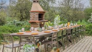 Area pranzo all'aperto con forno a mattoni, sedie verdi e tavolo apparecchiato per un pasto.