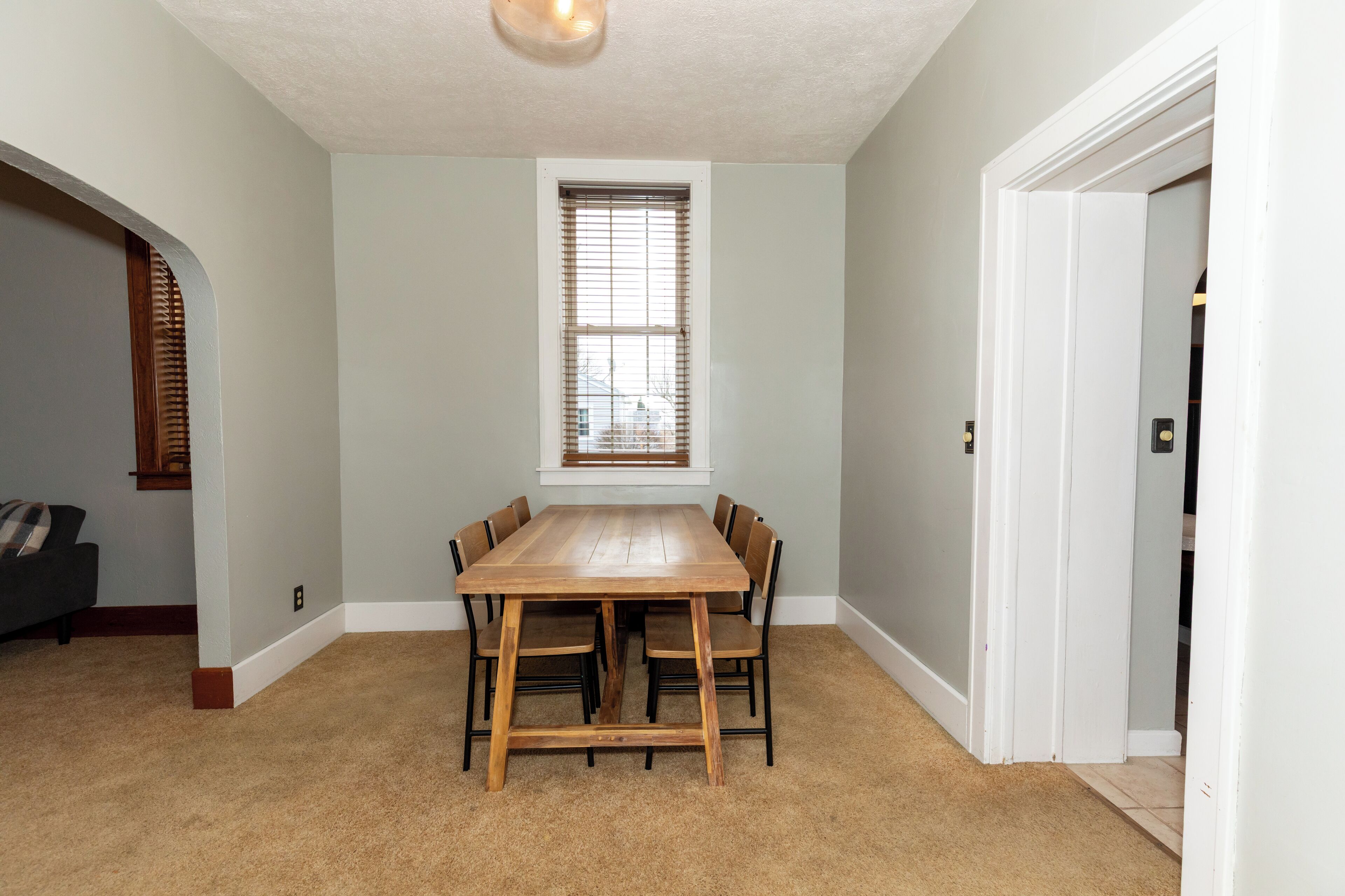 A dining area with a wooden table and chairs, a window with blinds, and a carpeted floor.