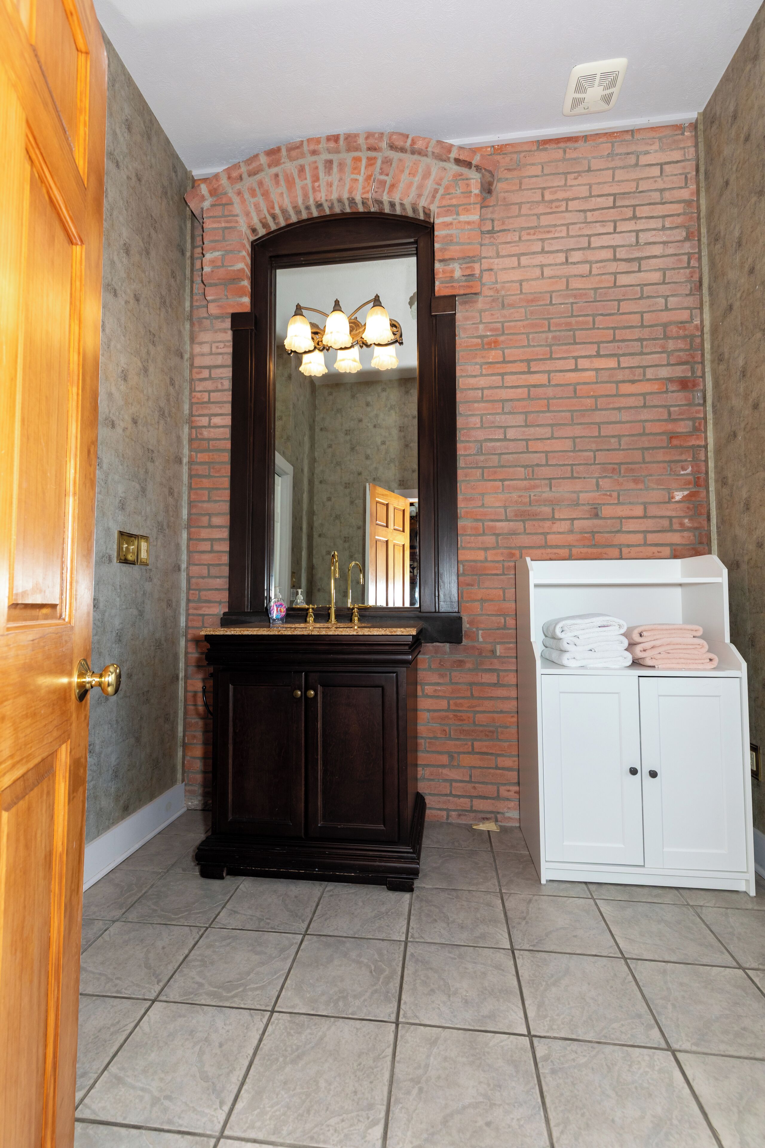 A bathroom with a wooden door, a dark wooden vanity with a mirror, a white cabinet with towels, and a brick wall.
