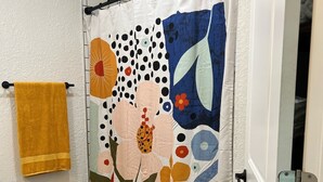 A bathroom with a shower curtain featuring a floral pattern, a yellow towel, a wicker basket, and a yellow rug. - Updated Bungalow - Close to town and 30 min from Lubbock! (Brownfield)