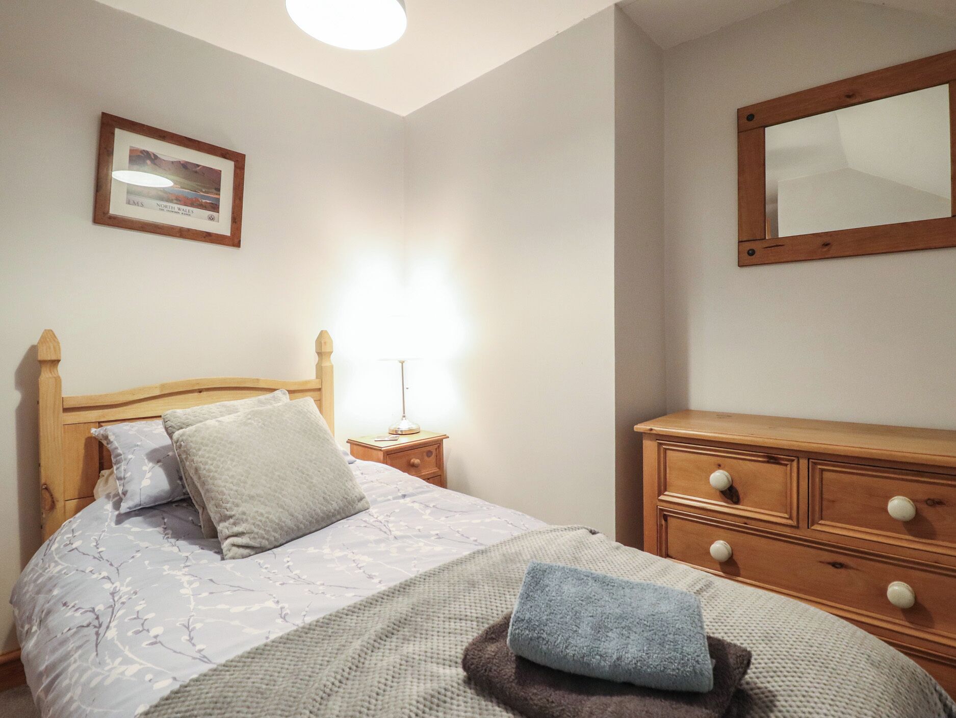 A bedroom with a wooden bed, a nightstand with a lamp, a dresser, and a framed picture on the wall.