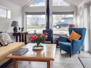 Un salón acogedor con un sofá, un sillón azul, una mesa de centro con un libro y un jarrón con flores. Se ve una chimenea a través de la ventana, y un coche aparcado fuera.