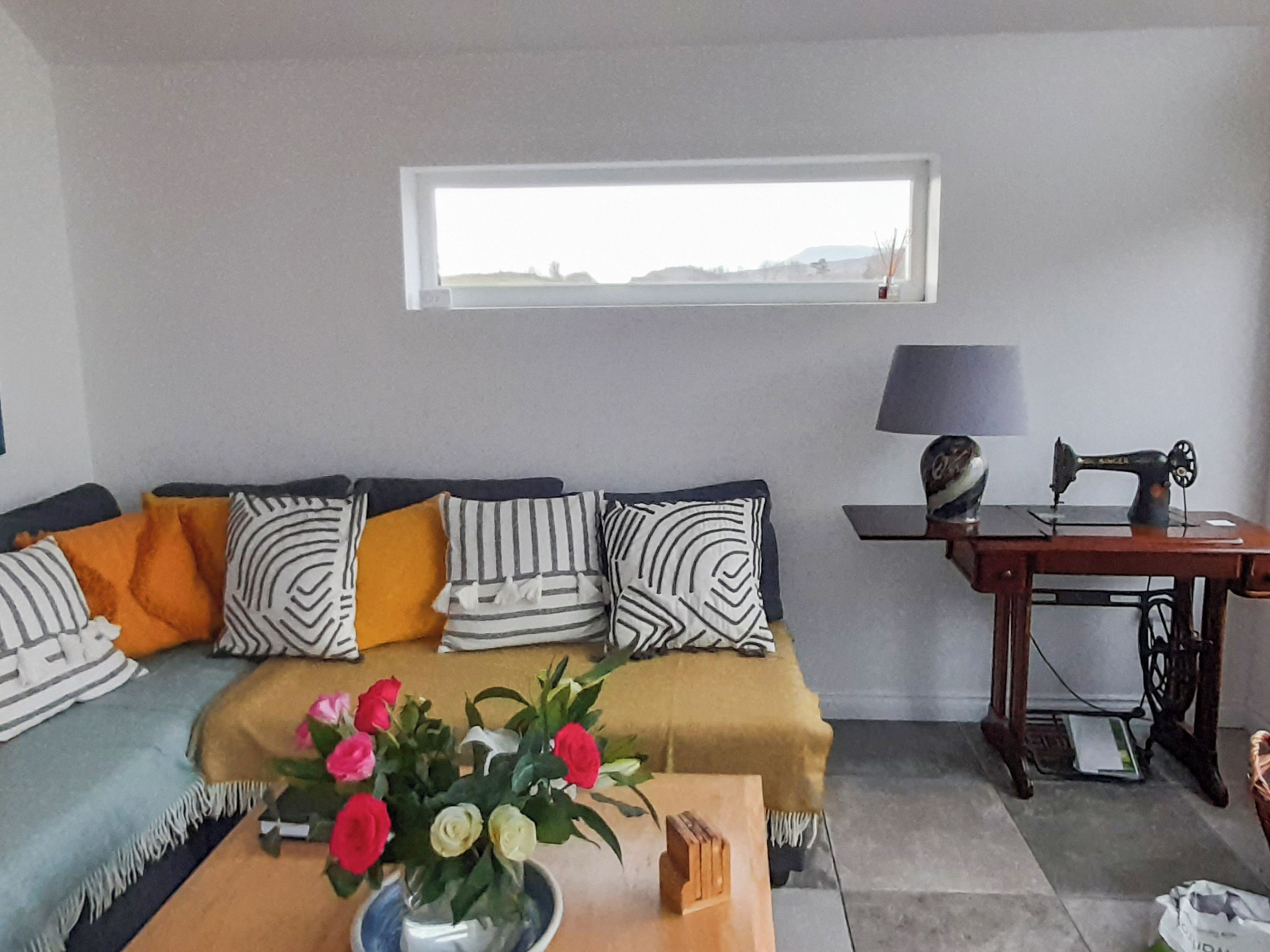 A living room with a sofa, a table with a vase of flowers, a sewing machine, and a window with a view.