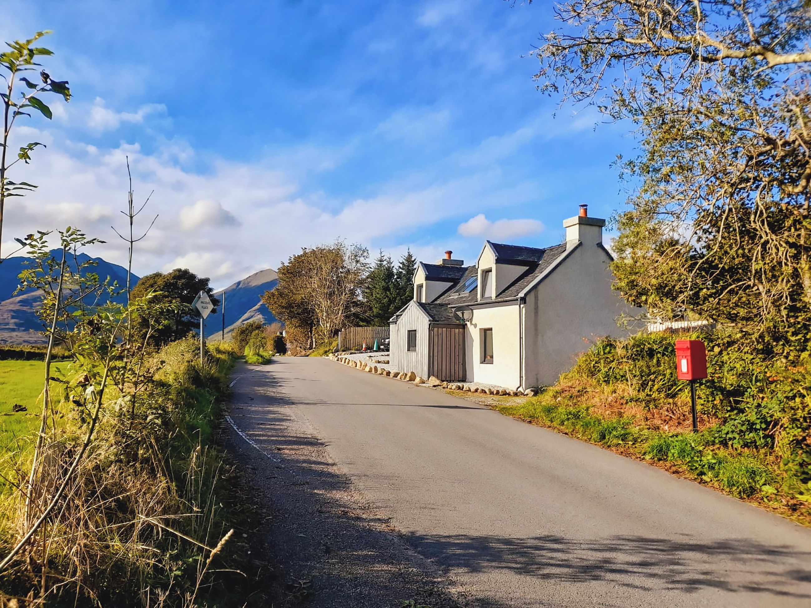 A rural road with a white house on the right, surrounded by greenery and mountains in the background.
