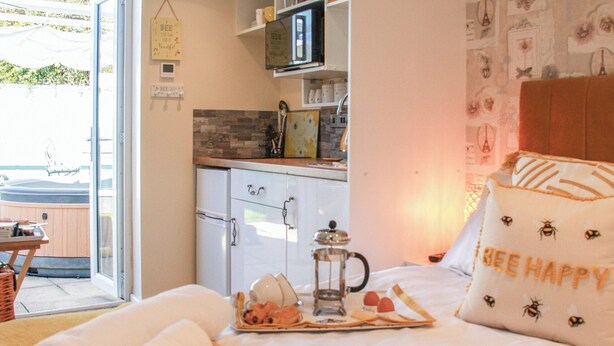 A cozy bedroom with a bed, a bedside table with a tray of breakfast, a window with a view of a hot tub, and a kitchenette in the background. - 1 bedroom accommodation in Mundesley (Mundesley)