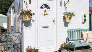 A white building with a door marked "WELCOME," two hanging flower baskets, a bench with a cushion, and a small table with a cup and saucer. - 1 bedroom accommodation in Mundesley (Mundesley)