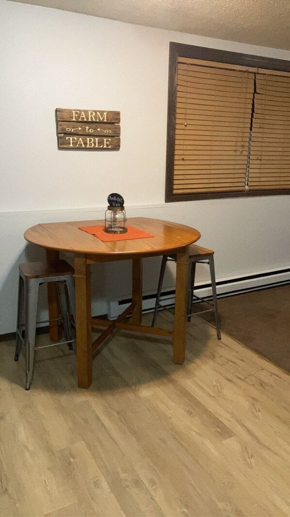 A round wooden dining table with two metal stools, a wooden sign above reading "FARM to TABLE", and a window with blinds in the background. - HC Hideaway - Rustic Roomy Relaxing! Queen Bed, Full Kitchen! (Aberdeen)