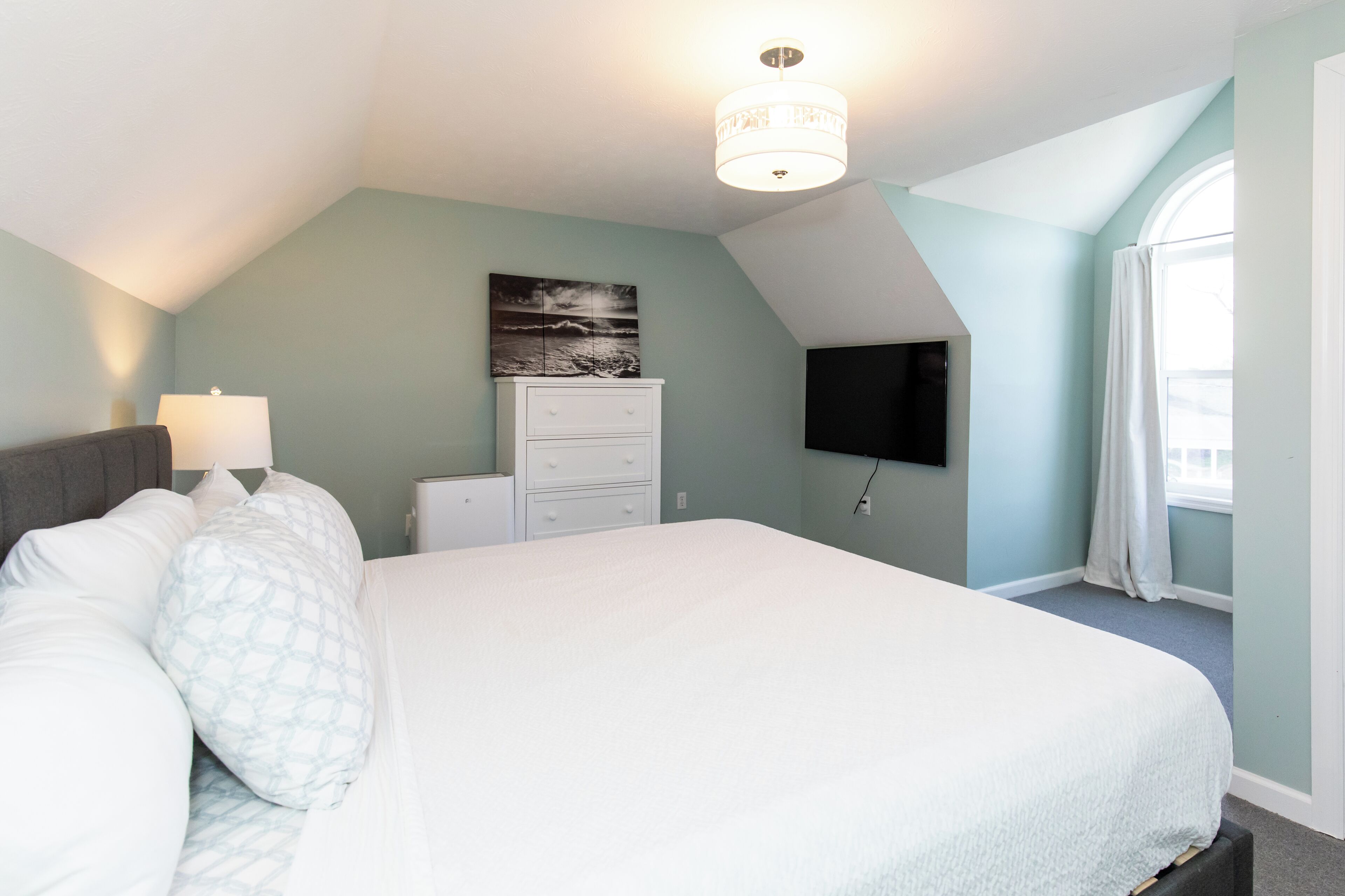 A bedroom with a large bed, a dresser, a flat-screen TV mounted on the wall, and a window with curtains.