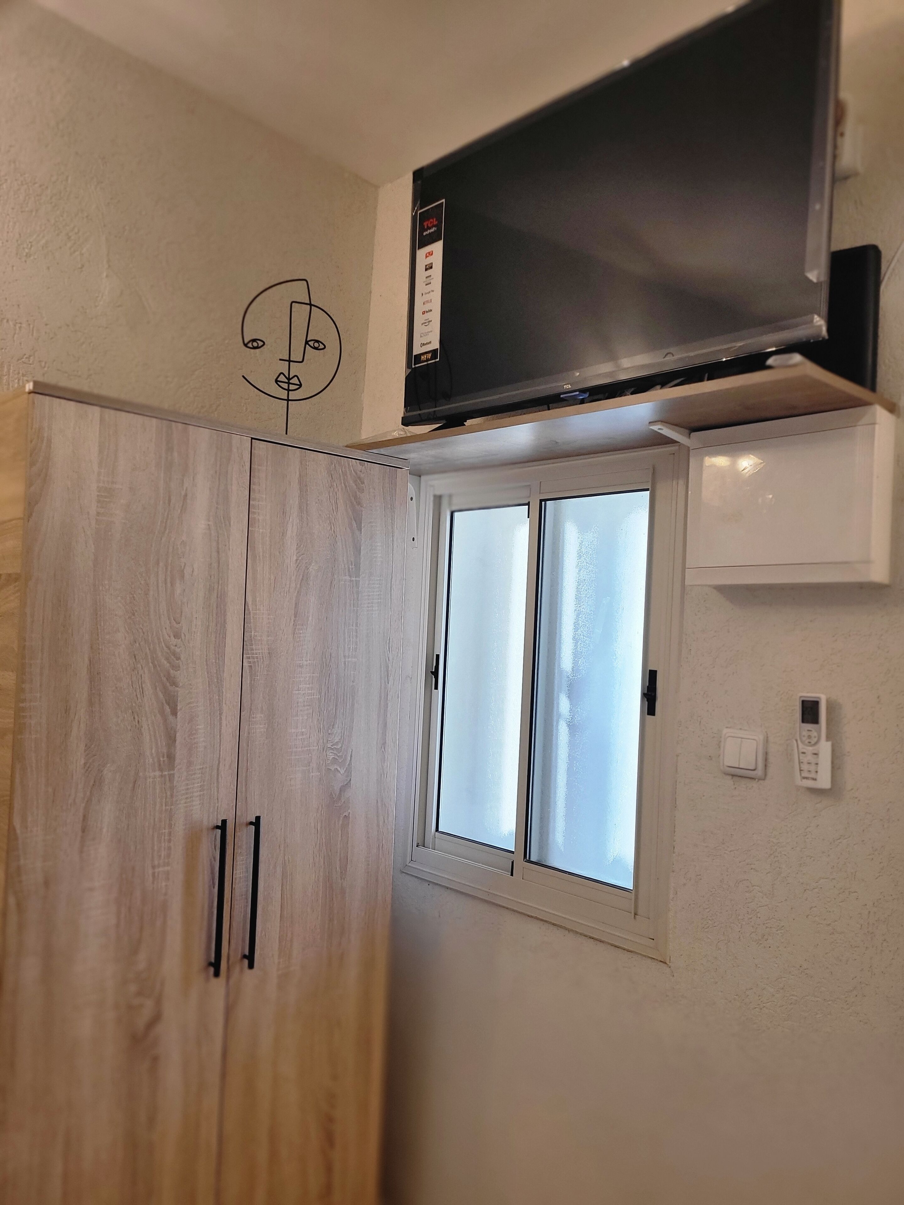 A room with a wooden wardrobe, a window with frosted glass, a wall-mounted TV, and a thermostat.