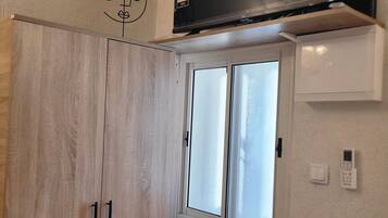 A room with a wooden wardrobe, a window with frosted glass, a wall-mounted TV, and a thermostat.