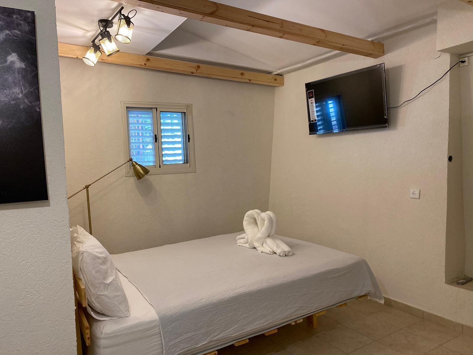 Um quarto pequeno e bem arrumado, com uma cama de solteiro, uma janela, uma TV montada na parede e uma luminária.