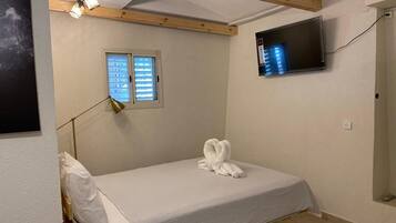 Um quarto pequeno e bem arrumado, com uma cama de solteiro, uma janela, uma TV montada na parede e uma luminária.