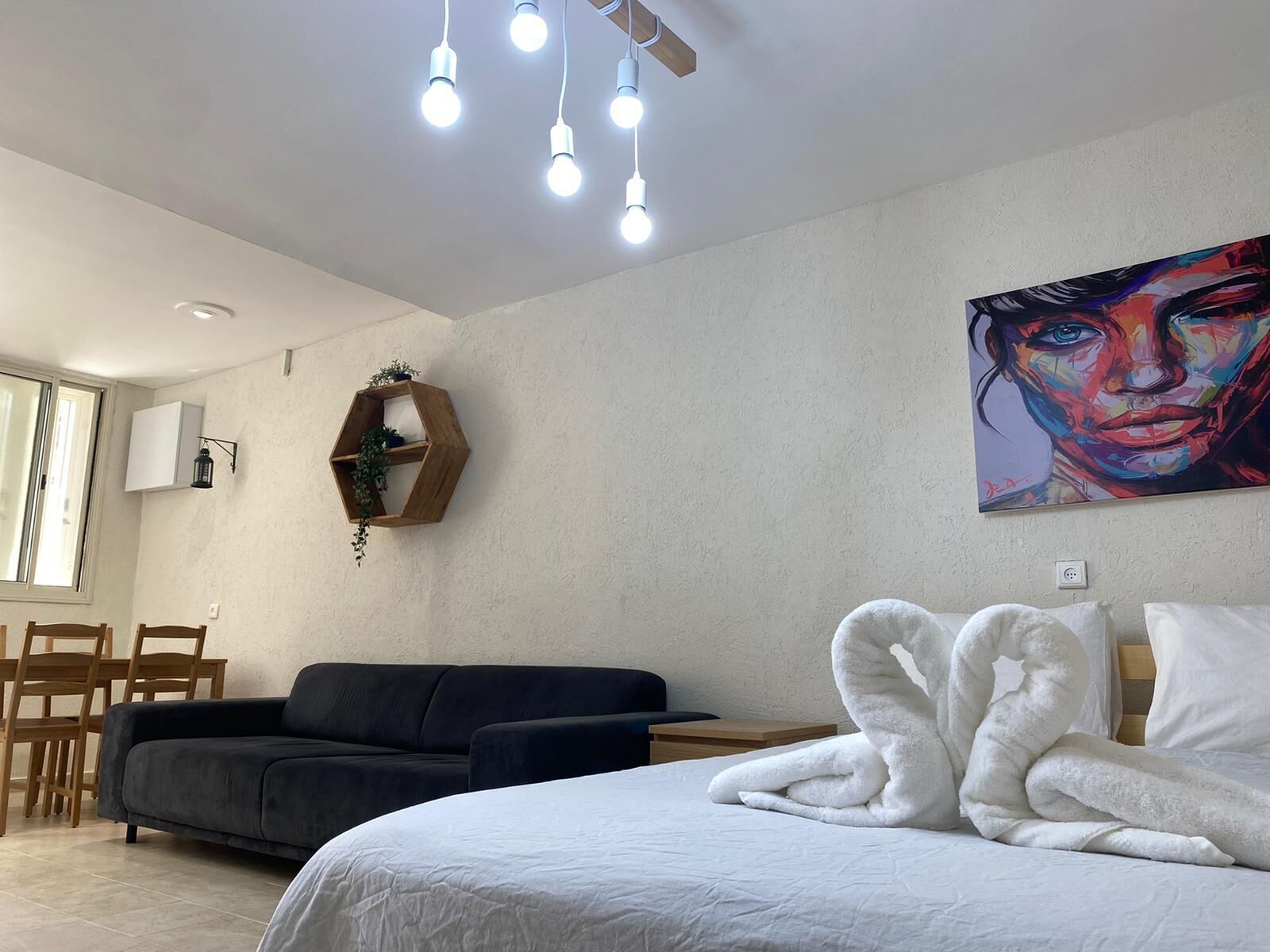 Une chambre à coucher moderne avec un lit, un canapé foncé, une étagère en bois avec des plantes, un tableau et un luminaire au plafond.