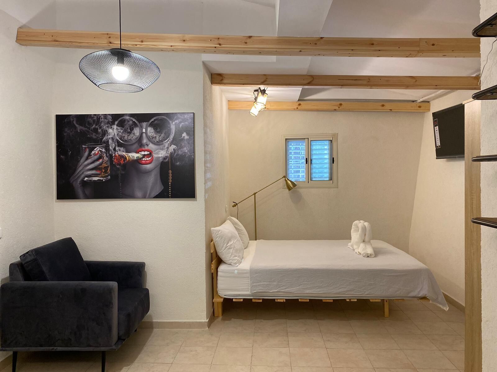 A compact bedroom with a bed, a black chair, a painting, a window, and a ceiling light.