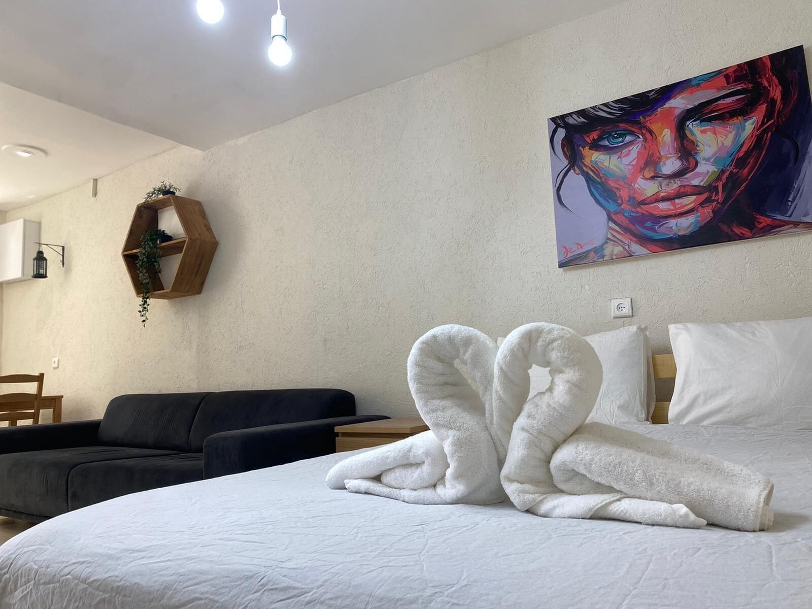 Une chambre d’hôtel avec un lit, un tableau au mur, une étagère en bois avec une plante et un canapé noir.
