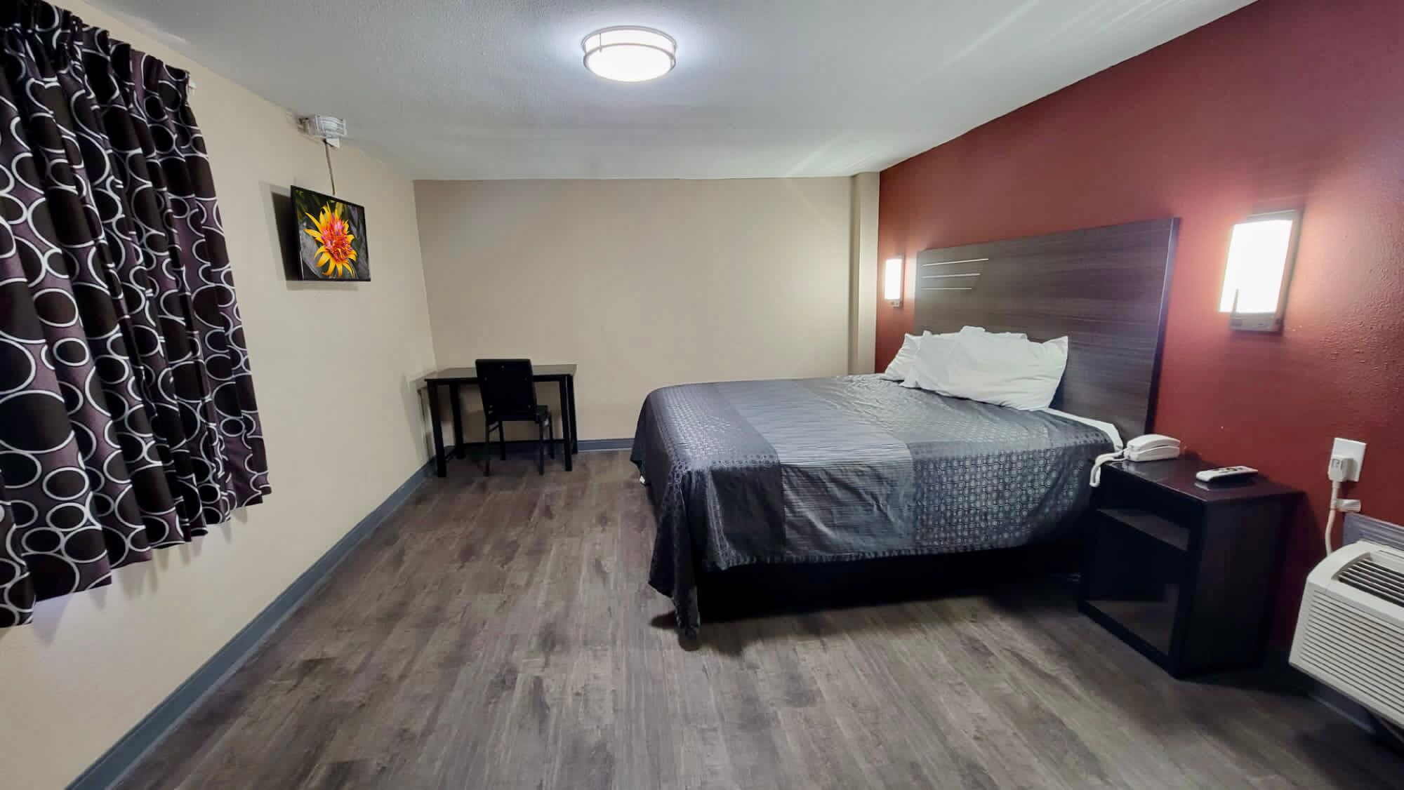 A hotel room with a bed, a desk, a chair, a wall-mounted air conditioner, and a ceiling light.