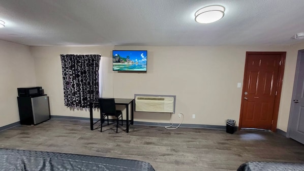 Una habitación pequeña y amoblada con escritorio, silla y refrigerador. Se observa un cuadro en la pared, una puerta y un bote de basura.