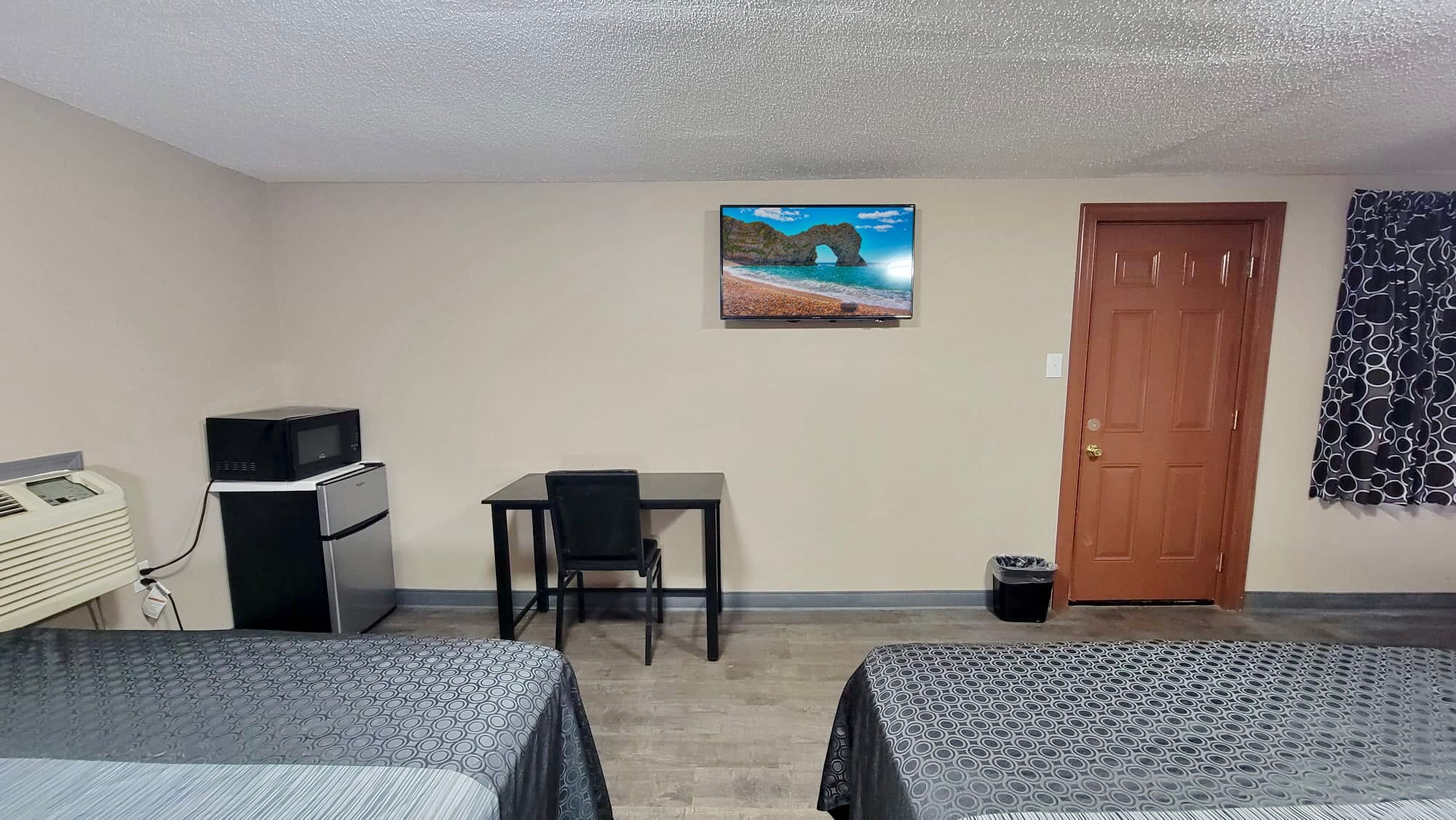 A hotel room with two beds, a desk, a television, a refrigerator, a microwave, and a door.