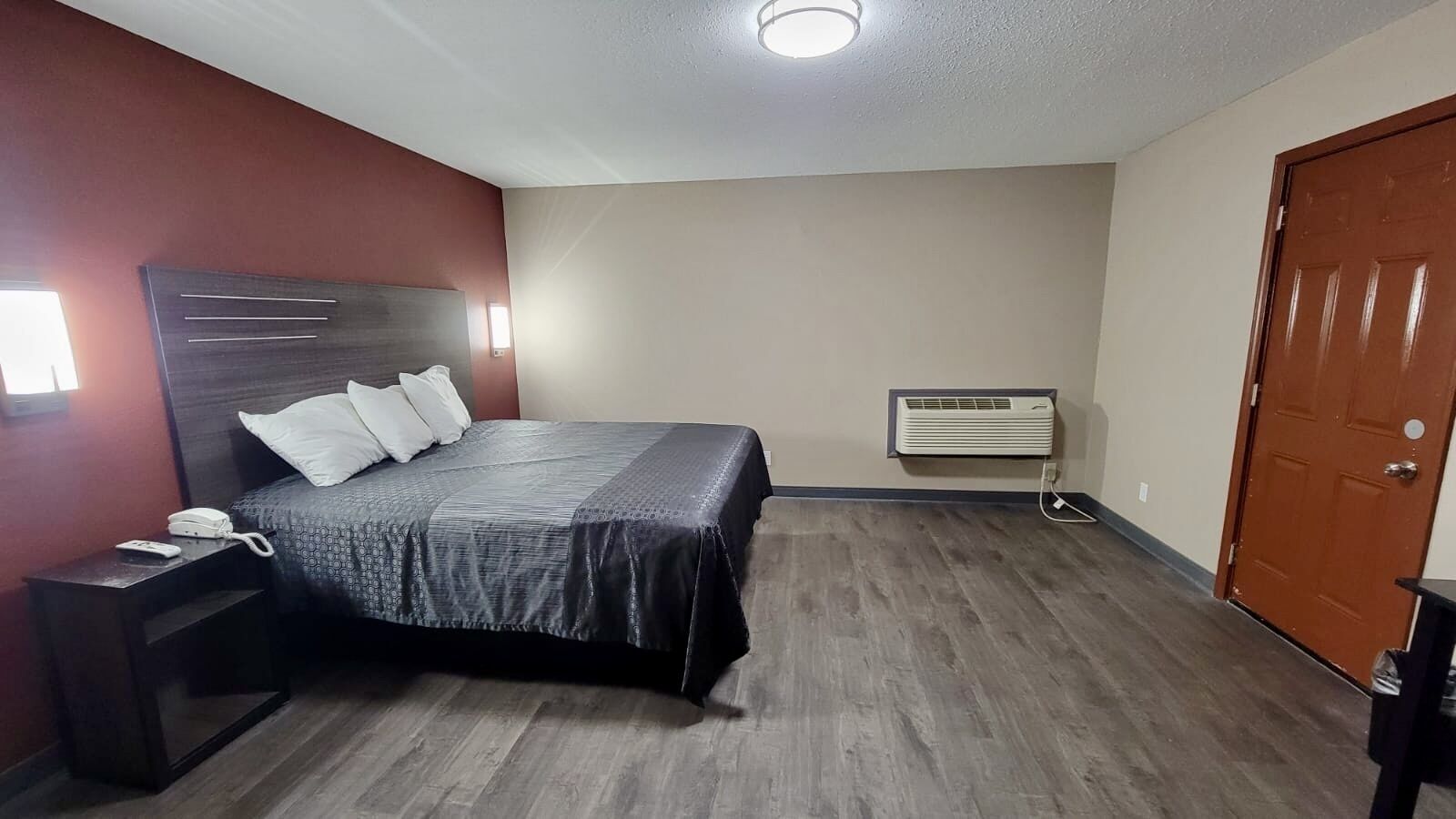 A hotel room with a bed, a bedside table with a phone, a wall-mounted air conditioner, and a door.